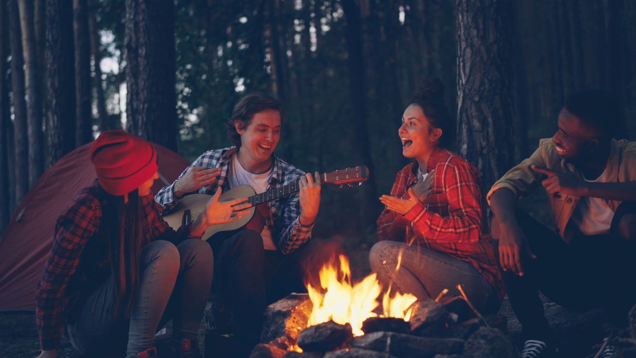 Four friends enjoy playing guitar and singing around a campfire in the woods.