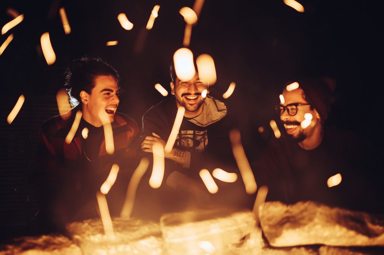 Three friends laughing and enjoying a bonfire at night, surrounded by warm firelight.