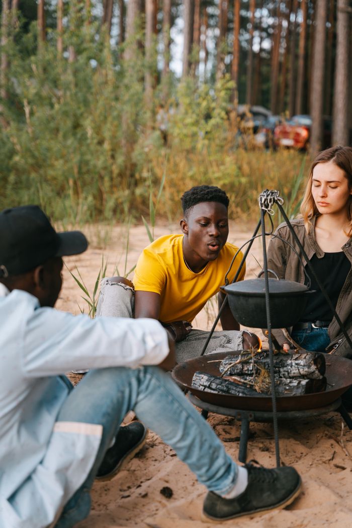 A group of young adults gather around a campfire, cooking and enjoying nature outdoors.