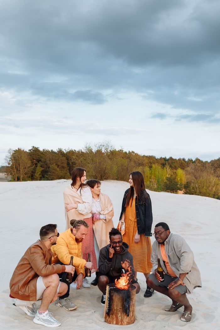 Group of friends having fun around a beach bonfire under cloudy skies.